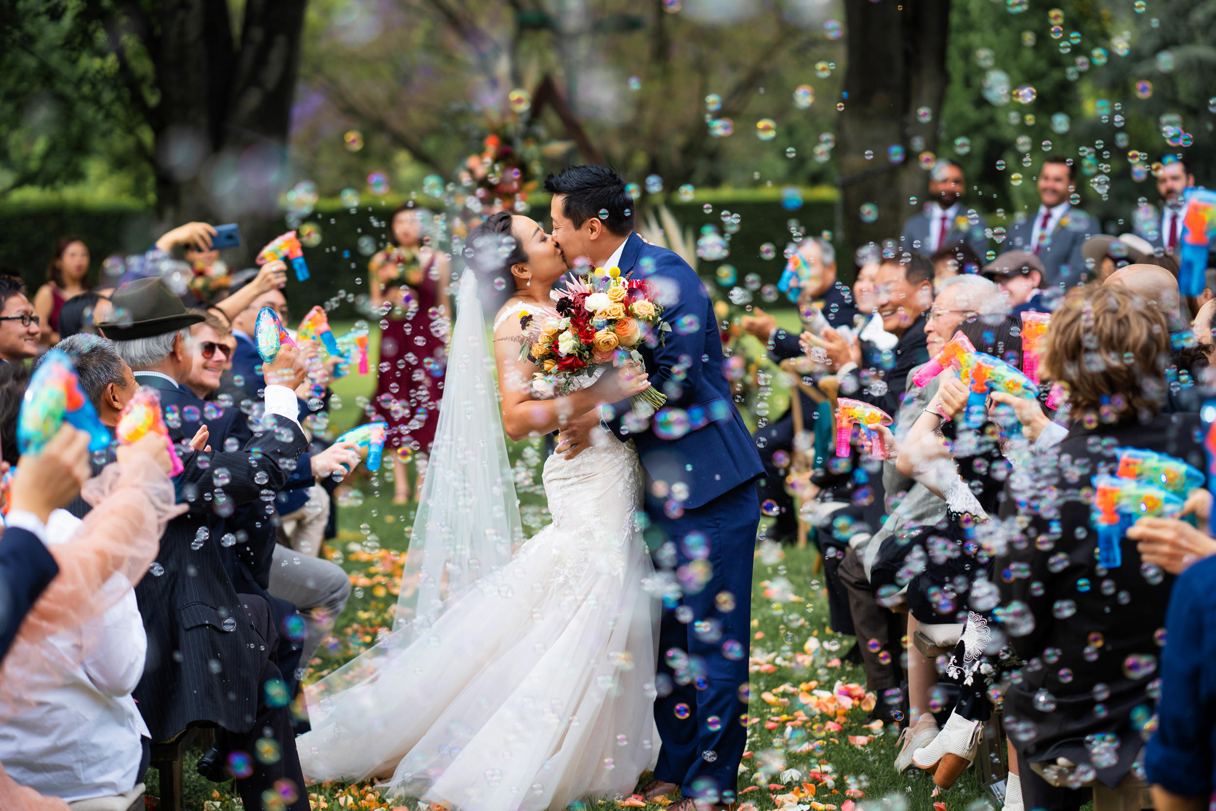 Bride and groom kissing with guests showering them with bubbles after their garden ceremony.