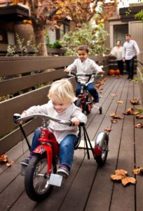 Two young boys race away from parent on red tricycles at their home.
