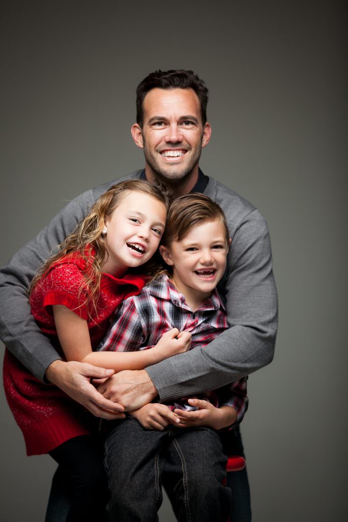 Single dad hugging young kids in portrait studio with grey backdrop.