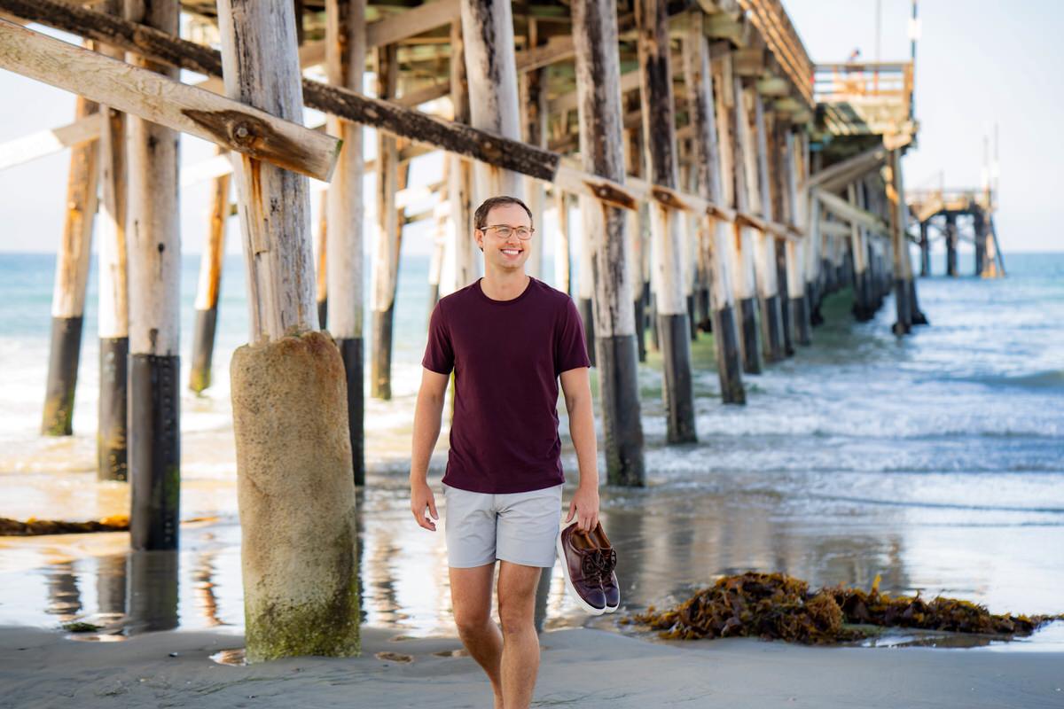 An authentic outdoor photo shoot features a young man walking on the beach in a distinctive burgundy t-shirt for his dating app profile.