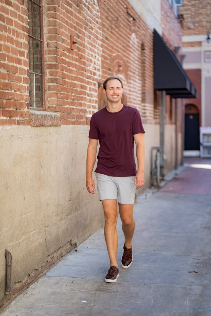 In his dating app profile image, a young man strolls along the beach, radiating a laid-back vibe in a fashionable burgundy t-shirt