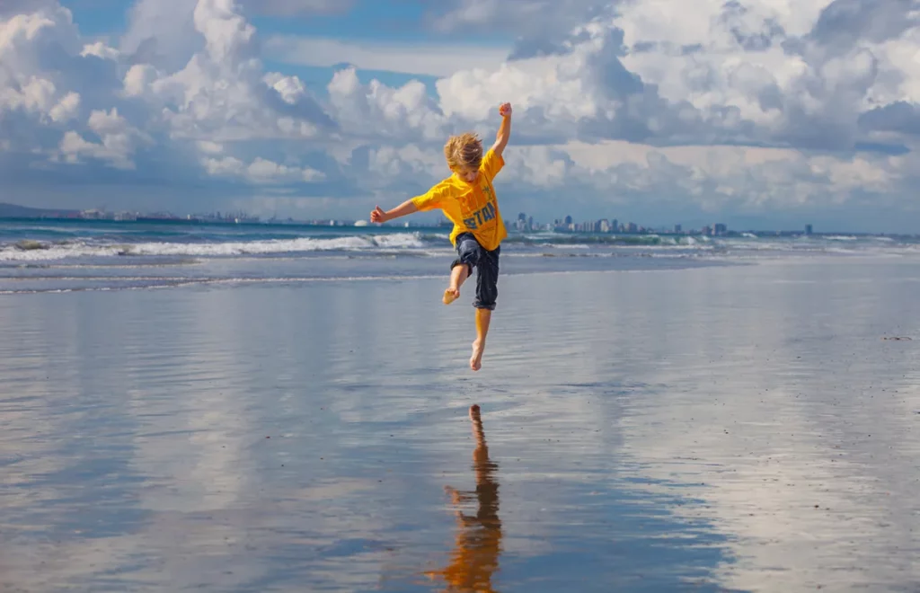 small boy wearing yellow shirt jumping during low tide looking at reflection in sand