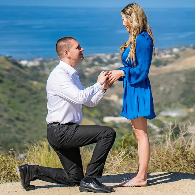 Guy down on one knee proposing to girlfriend at overlook location with ocean views.