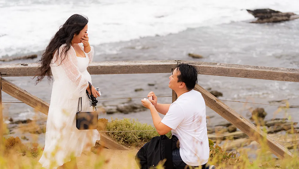Man proposing on one knee at beach to surprised girlfriend wearing white dress