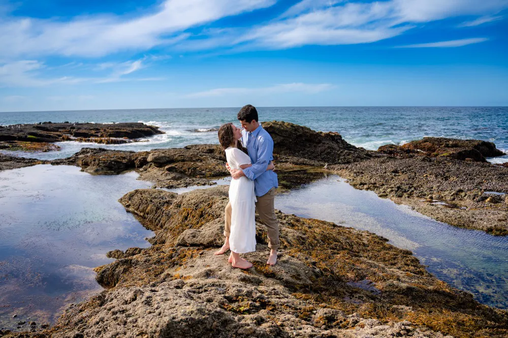 couple standing on rocks at beach during engagement photos