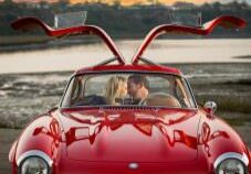 Engaged couple kiss in classic red mercedes at sunset.