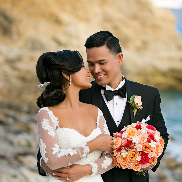 Bride adn Groom embrace on beach holding wedding bouquet